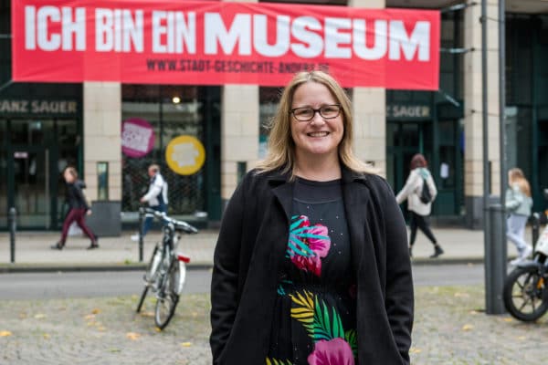 Das Bild zeigt die stellv. Museumsleiterin Silvia Rückert vor dem Stadtmuseum. Im Hintergrund ist ein roter Banner an der Fassade zu sehen mit der Aufschrift "Ich bin ein Museum".