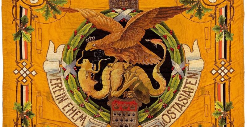 Man sieht eine Fahne. Diese ist gelb. Darauf zu sehen sind mehrere Blumenornamente sowie ein Adler der mit einem Drachen kämpft.