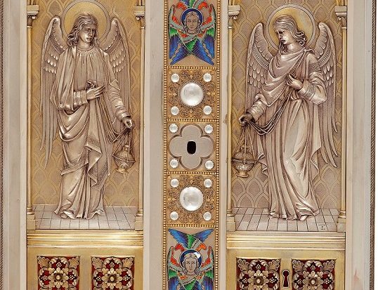 Zu sehen ist eine Tabernakeltür. Man sieht zwei Engel, einer auf der linken und einer auf der rechten Seite. Die Verzierungen sind aus Gold.