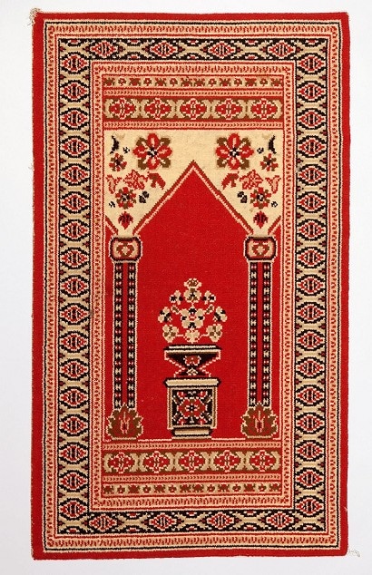 Man sieht einen Gebetsteppich mit einem orientalischen Muster. Die Farbe rot dominiert.