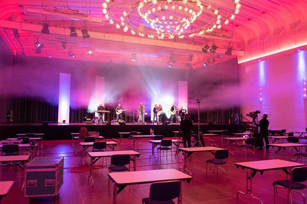 Zu sehen ist eine Bühne mit Musikern. Der Konzertraum erstrahlt in pinkem Licht.
