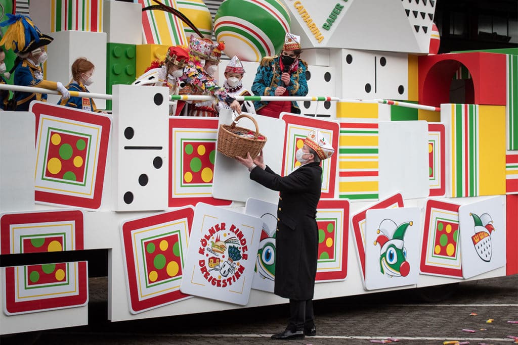 Man sieht einen Karnevalisten, wie er an einem Karnevalswagen steht. Er bekommt einen Korb mit Kamelle.