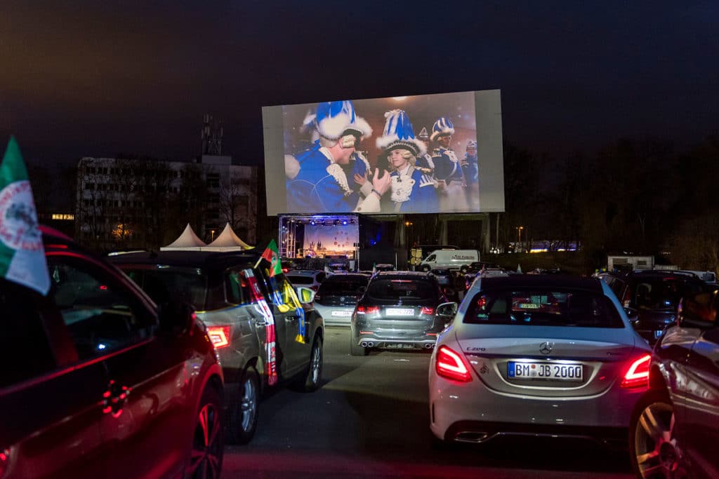 Zu sehen ist ein Foto der Veranstaltung "Alaaf auf Abstand". Man sieht Autos bei Nacht und eine Leinwand, auf der die Karnevals-Veranstaltung übertragen wird.
