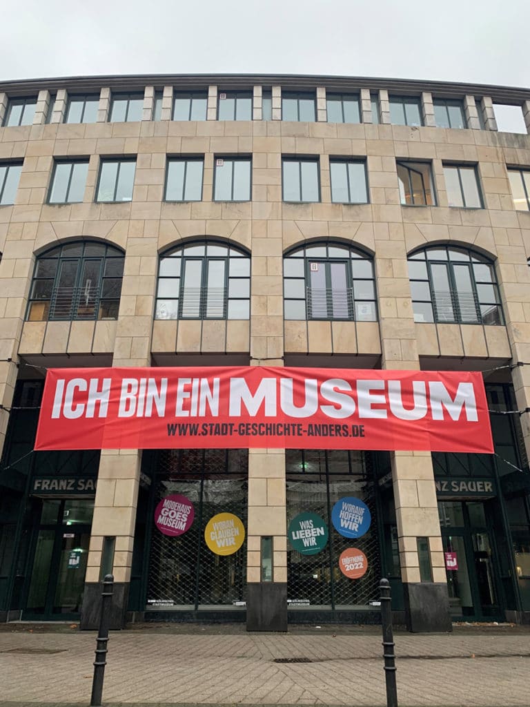 Man sieht die Außenfassade des neuen Museums mit einem roten Banner. Darauf steht "Ich bin ein Museum".