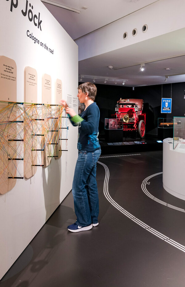 Besucherin an einer Mitmachwand mit bunten Fäden, dahinter ein historisches rotes Fahrzeug