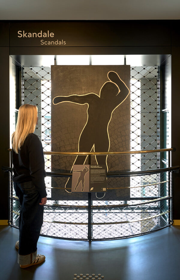 Besucherin vor einer beleuchteten Vitrine im Bereich „Skandale", darin eine goldfarbene Tatortsilhouette.