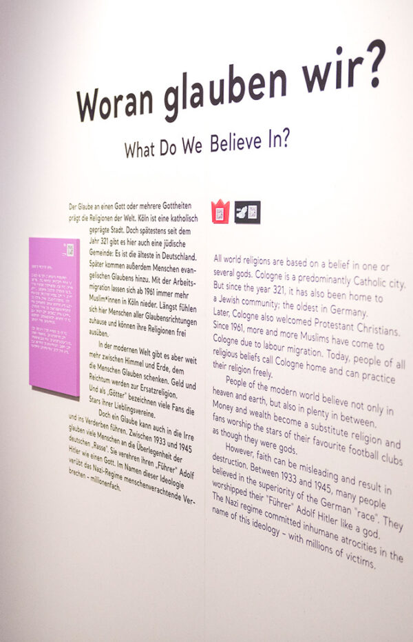 Wandtext der Ausstellung mit der Frage „Woran glauben wir?" auf Deutsch und Englisch